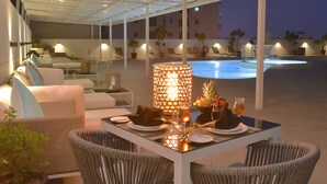 Ein Essbereich mit einem Tisch für zwei Personen, umgeben von Liegestühlen und einem Pool im Hintergrund.