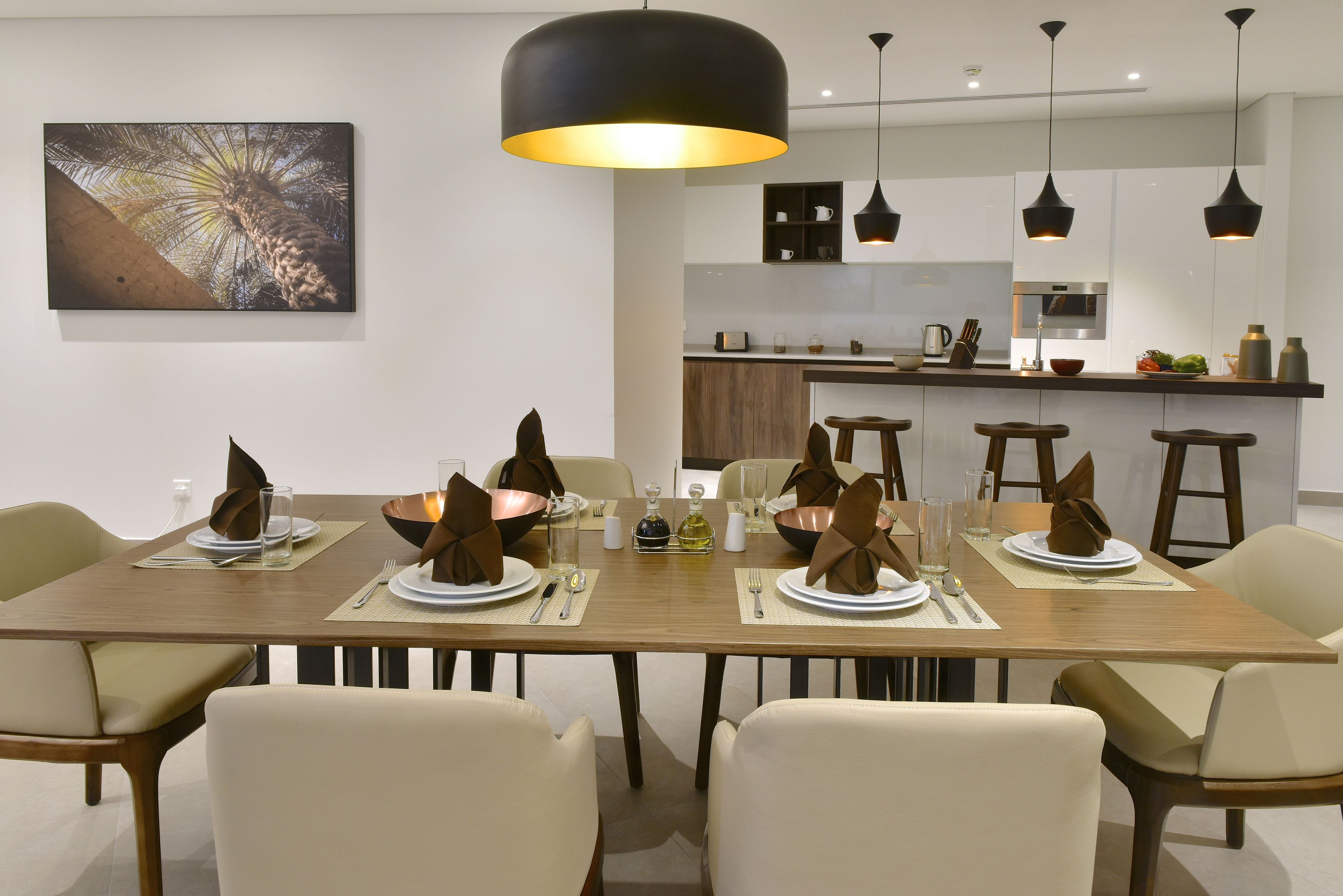 Un comedor moderno con una mesa de madera dispuesta para seis personas, rodeada de sillas color beige. Una lámpara colgante negra de gran tamaño ilumina el espacio, y se puede ver una cocina al fondo.
