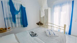 Ein ordentlich gemachtes Bett mit weißen Leinen, ein Nachttisch mit einer Fruchtkörbchen und einem Glas, sowie ein Fenster mit durchsichtigen Vorhängen.