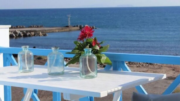 Ein weißer Tisch mit drei Glasflaschen und einer roten Blume auf einem blauen Geländer mit Blick aufs Meer.