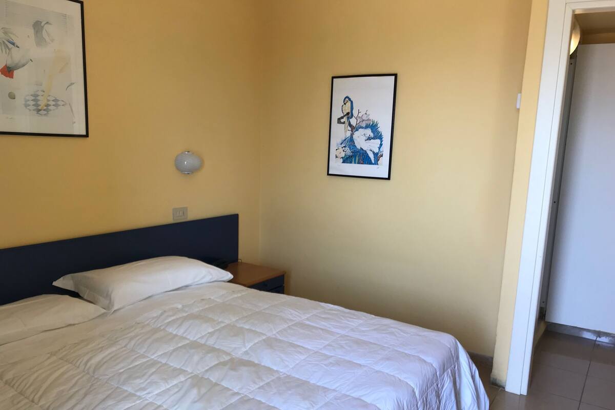 Een net opgemaakt bed met een witte quilt, een klein nachtkastje met een lamp, en twee kunstwerken in lijsten aan de muur.