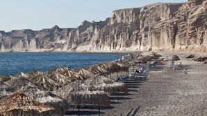 A beach with thatched umbrellas and sun loungers along the shore, cliffs in the background, and a clear blue sea. - 360 Blue (Santorini)