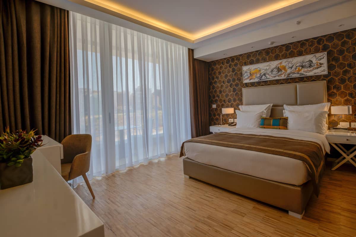 A modern hotel room with a large bed, a desk, and a view through sheer curtains.
