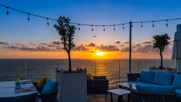 Ein Sitzbereich im Freien mit Tischen und Stühlen mit Blick auf einen Sonnenuntergang über dem Meer.