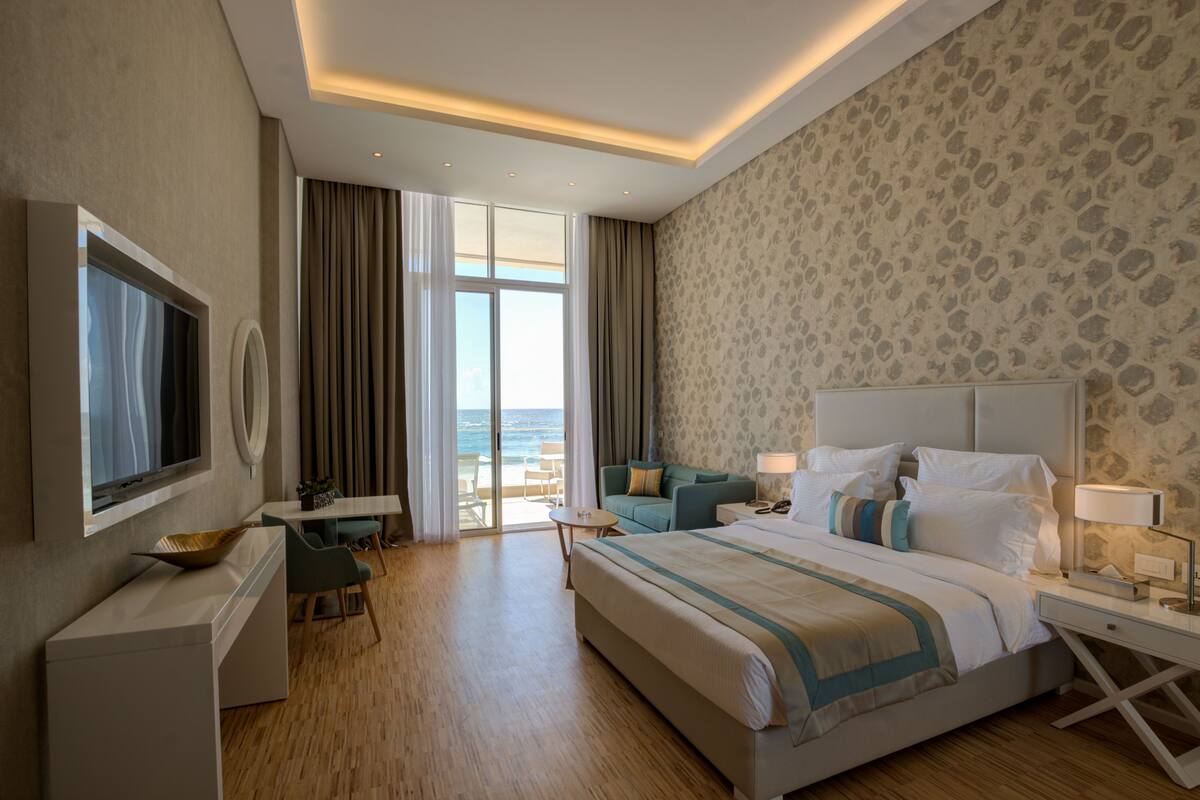 A hotel room with a bed, a desk, a TV, a dining area, and a view of the ocean.