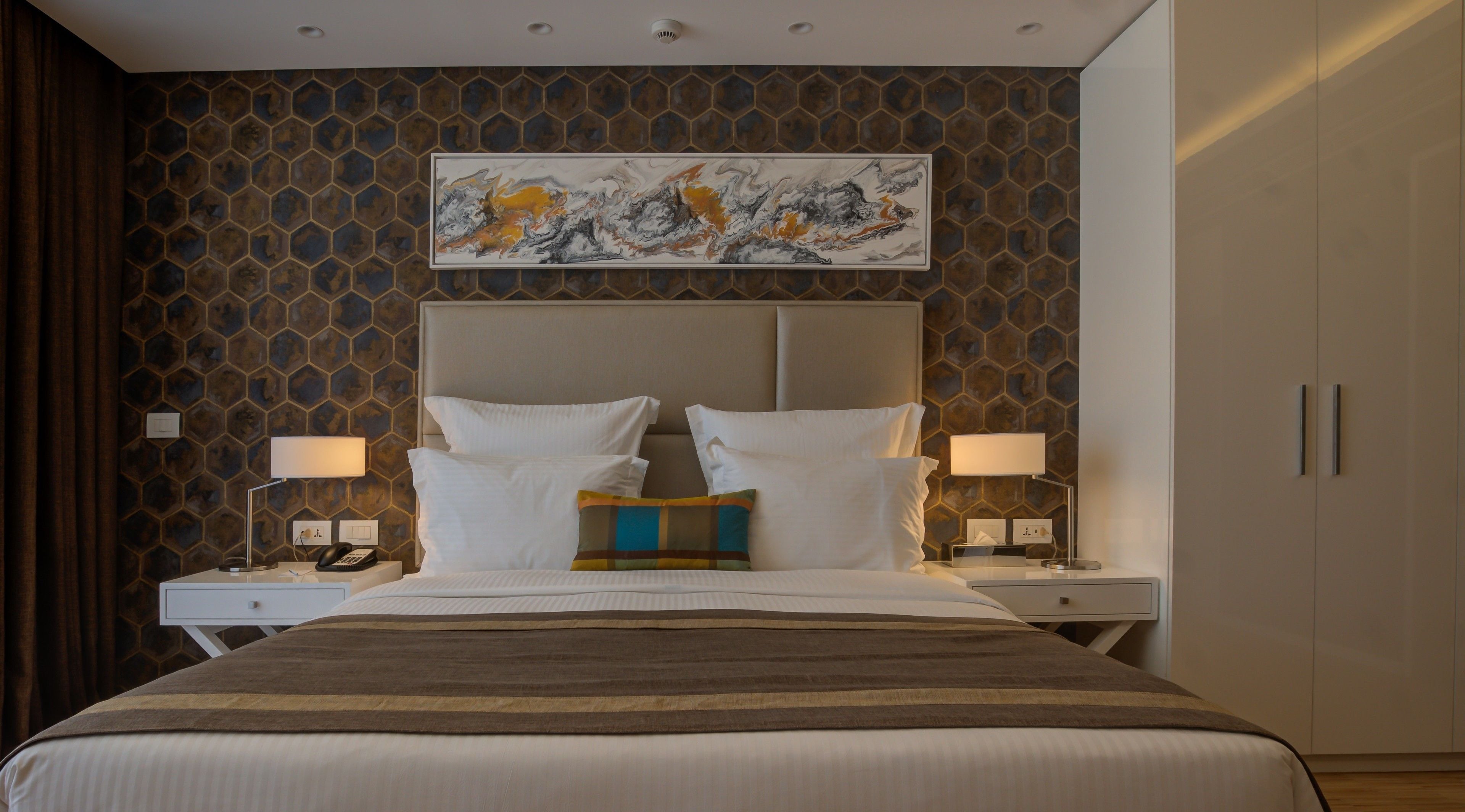 Habitación de hotel con una cama grande, dos mesitas de noche con lámparas, un cabecero y una obra de arte colgada en la pared.