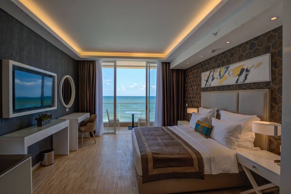 A modern hotel room with a large bed, a desk, a chair, and a view of the ocean.