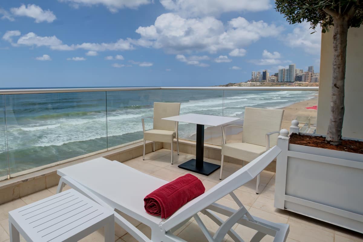 A seaside terrace with white furniture, a glass balustrade, and a city skyline in the background.