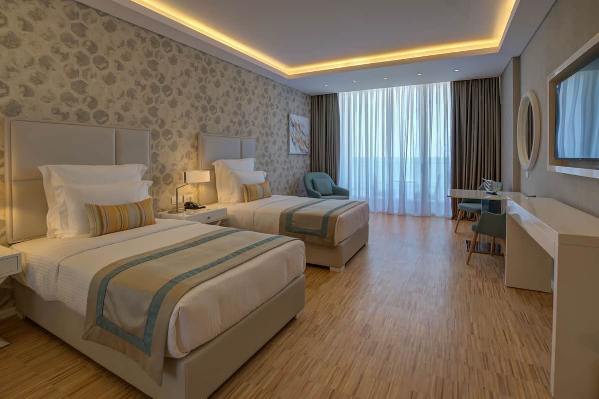 A hotel room with two beds, a desk, a chair, a TV, and a large window with curtains.