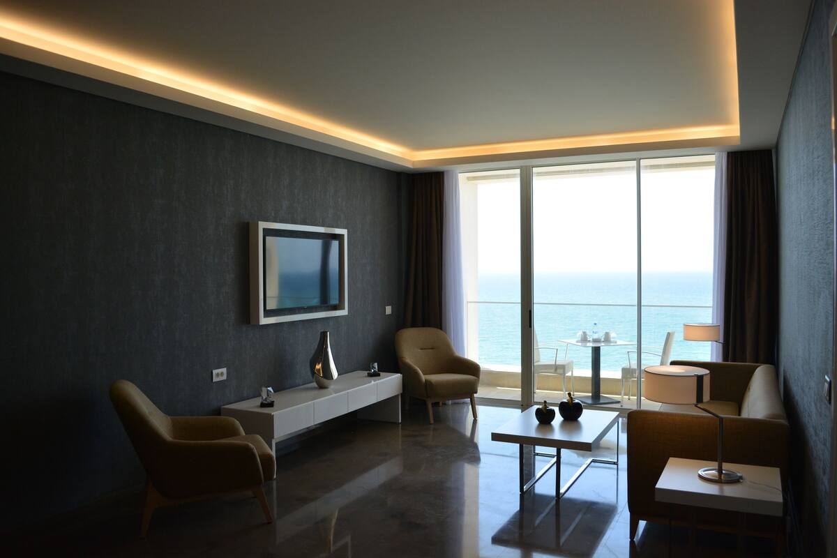 A modern hotel room with a large window, a flat-screen TV, a sofa, a coffee table, and a small table with a lamp.