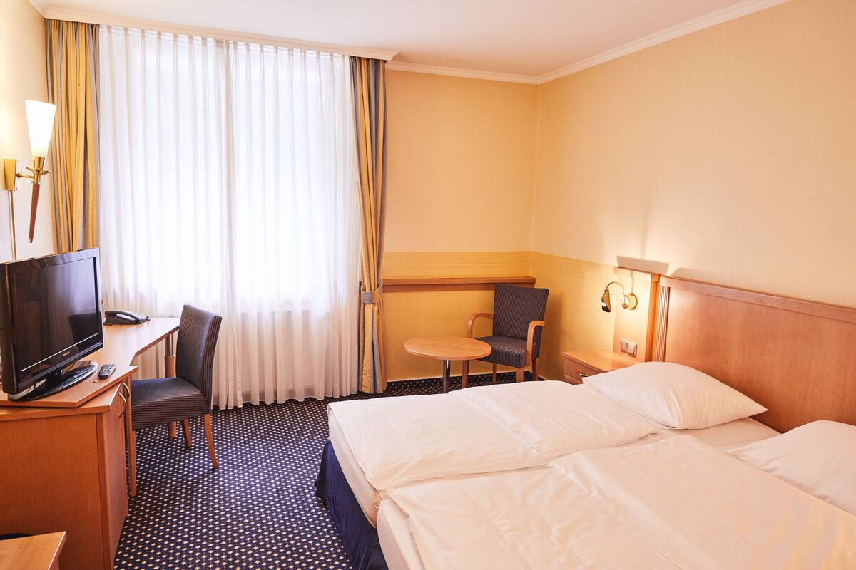 A hotel room with a bed, a desk with a TV and phone, a chair, a small table, and curtains.