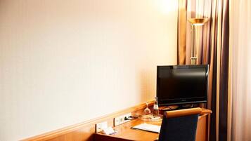 A hotel room with a wooden desk, a flat-screen TV, a glass of water, and a lamp.