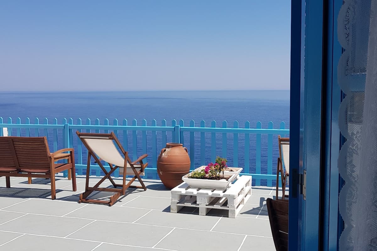 A balcony with a view of the sea, featuring wooden chairs, a table with a vase of flowers, and a large pot.