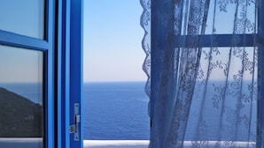 Une vue à travers une fenêtre à cadre bleu, ornée d’un rideau en dentelle, donnant sur un paysage côtier sous un ciel dégagé.