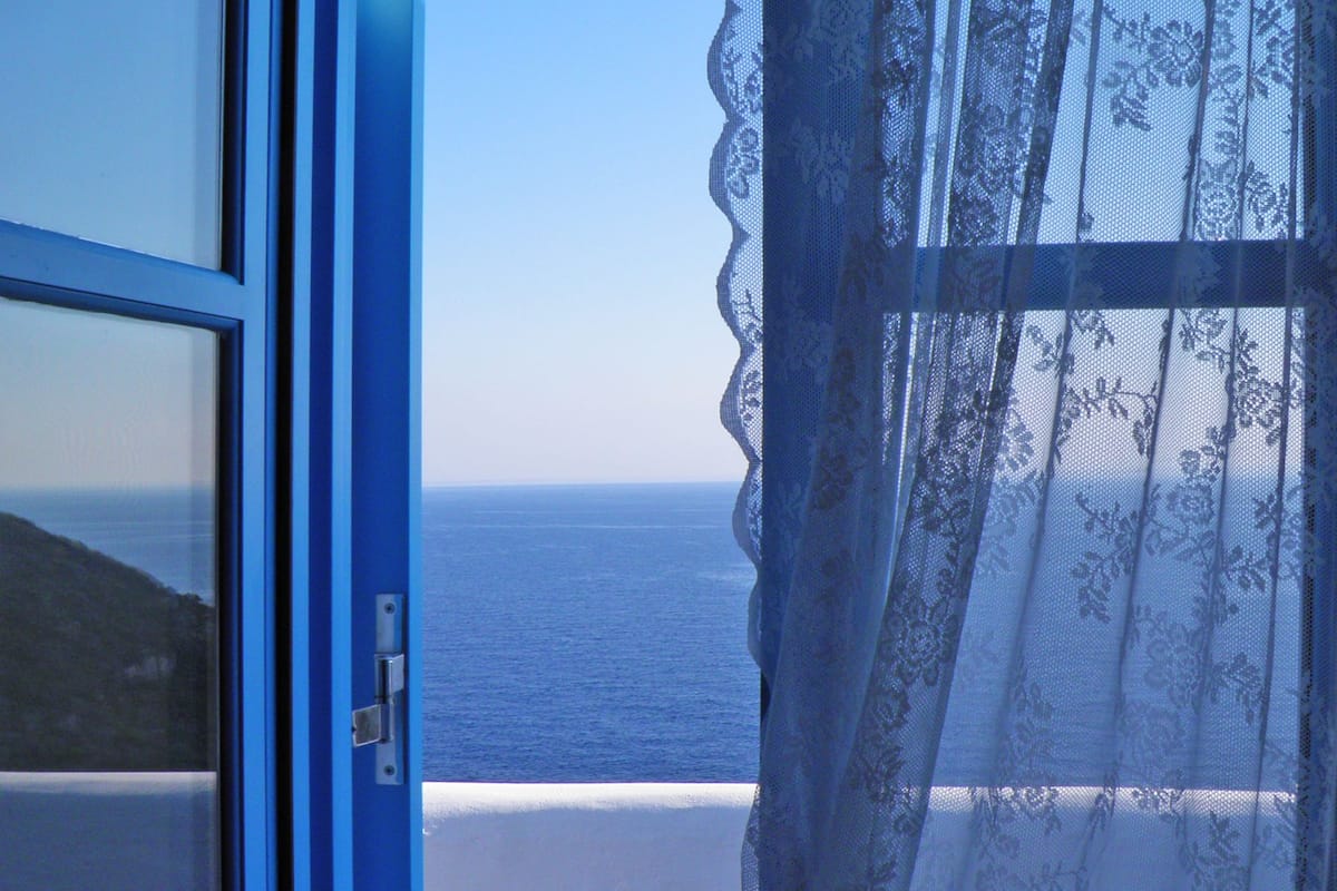 A view through a blue-framed window with a lace curtain, overlooking a coastal landscape with a clear sky.