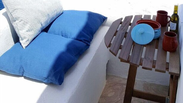 Un espace extérieur aménagé avec des sièges blancs, une table en bois, des coussins bleus et une bouteille de vin.