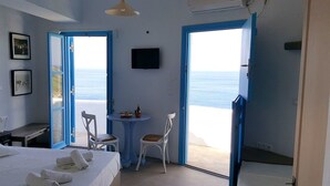 Une chambre avec un lit, une petite table et une chaise, un téléviseur, et une vue sur la mer par la porte.