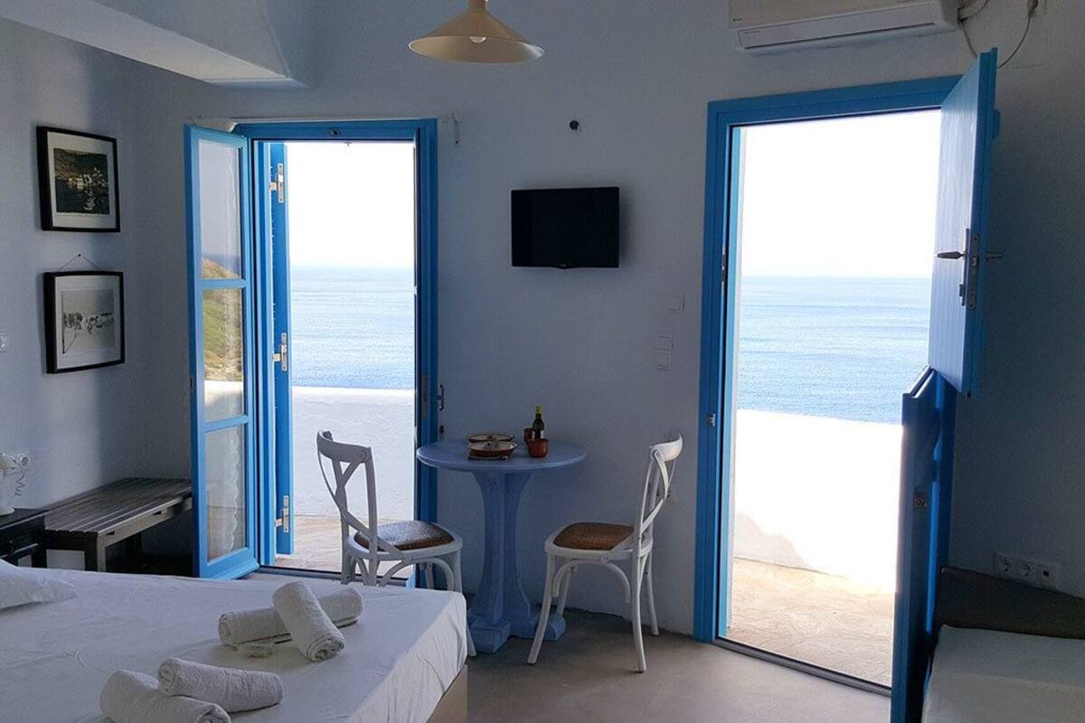 A room with a bed, a small table with a chair, a television, and a view of the sea through a door.