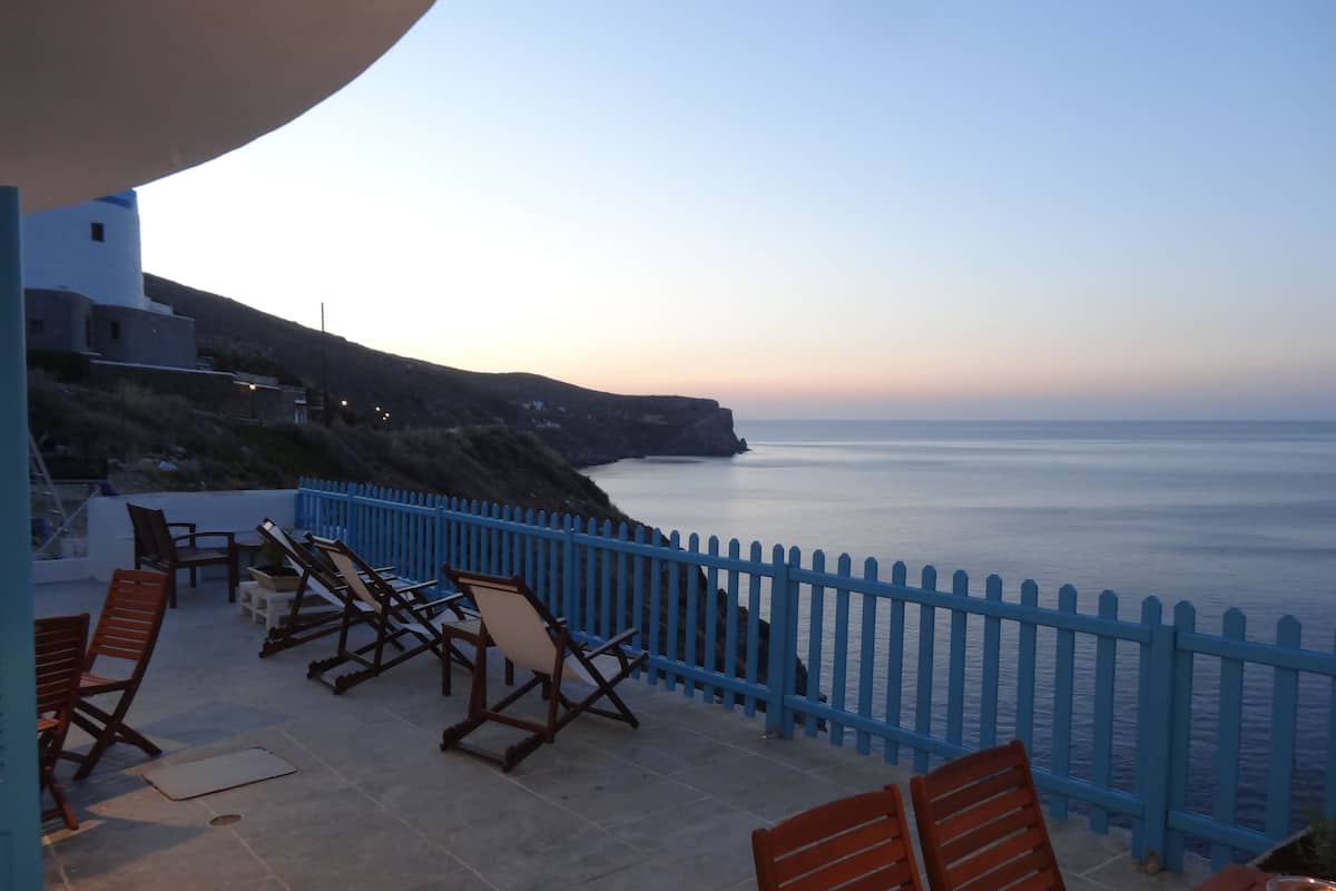 A terrace with chairs and a table, overlooking a coastal view with a blue fence and a cliff.