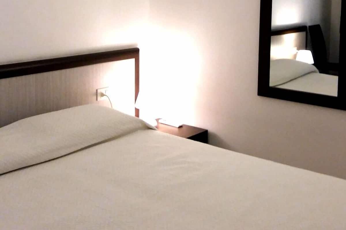 Een net opgemaakt bed met een witte dekbedovertrek en kussens, een nachtkastje met een lamp en een spiegel aan de muur.