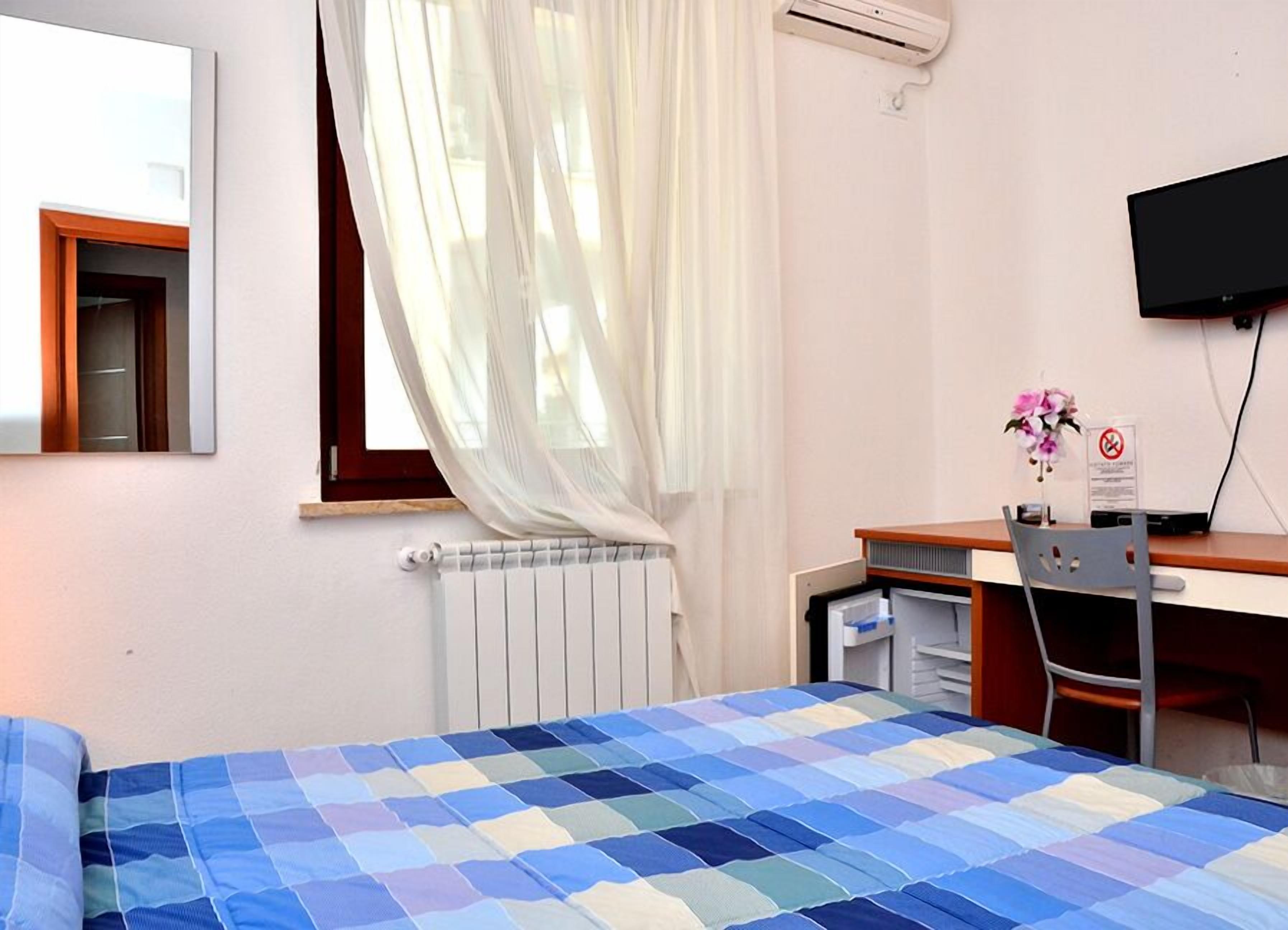 Une chambre à coucher comprenant un lit, un bureau, une chaise, un radiateur, une fenêtre avec des rideaux légers et un téléviseur à écran plat fixé au mur.