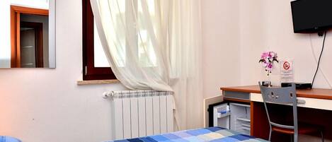 Une chambre à coucher comprenant un lit, un bureau, une chaise, un radiateur, une fenêtre avec des rideaux légers et un téléviseur à écran plat fixé au mur.