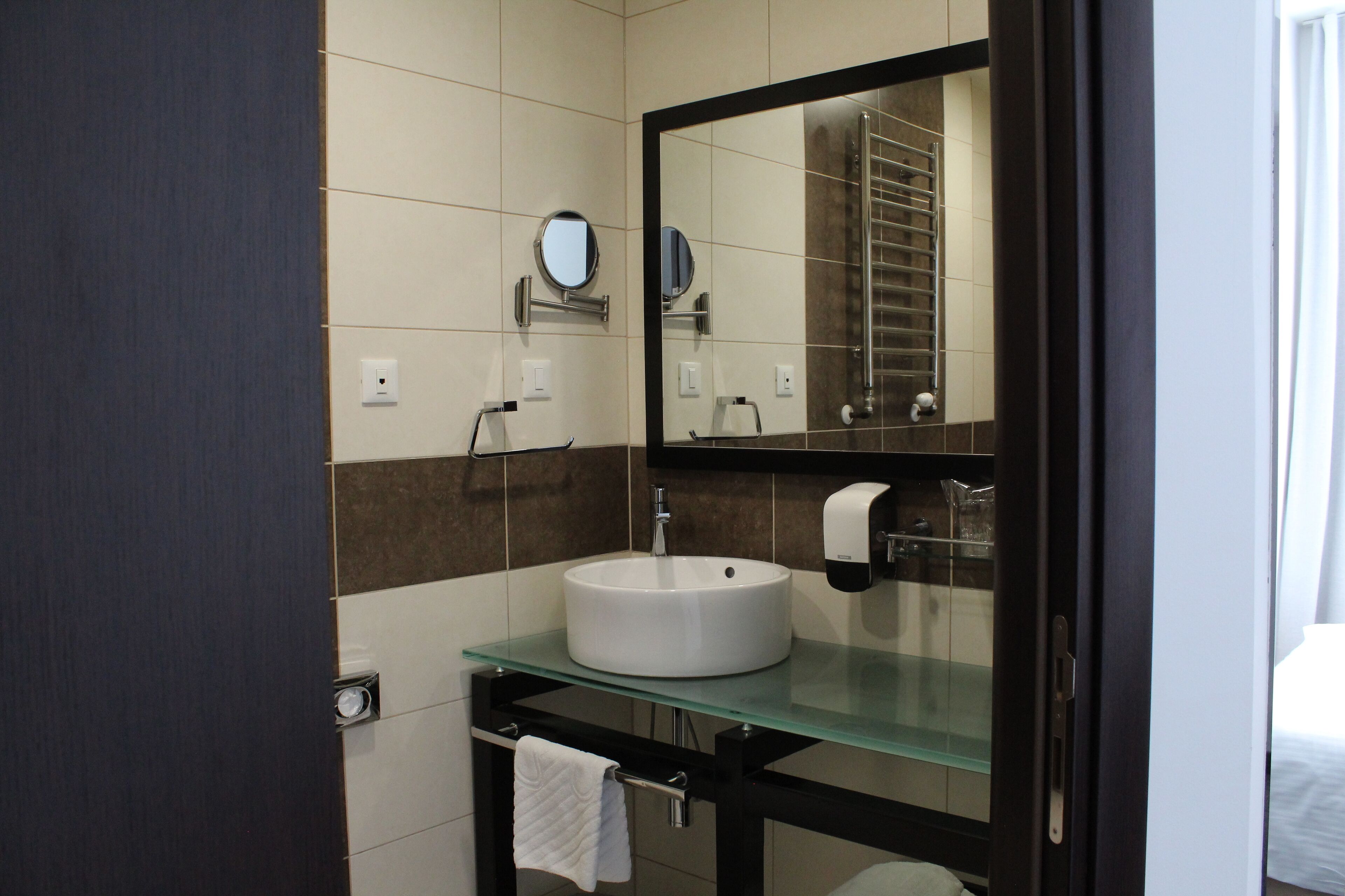 Un baño moderno con un lavabo blanco redondo, encimera de vidrio, un espejo grande y un perchero para toallas.