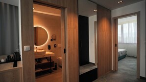 A modern hotel room with a wooden vanity, a round mirror, a sink, and a toilet. There is a bed with a white cover and a window with sheer curtains. - Hotel Diplomat (Zagreb)