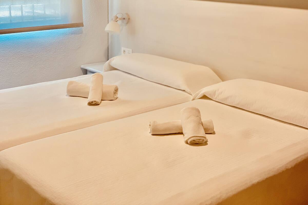 Ein Hotelzimmer mit zwei Betten, jeweils mit einem zusammengerollten Handtuch, einem Nachttisch und einer an der Wand befestigten Lampe.