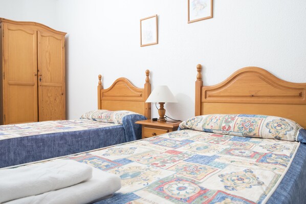 A bedroom with two beds, wooden headboards, a nightstand with a lamp, and a wardrobe.