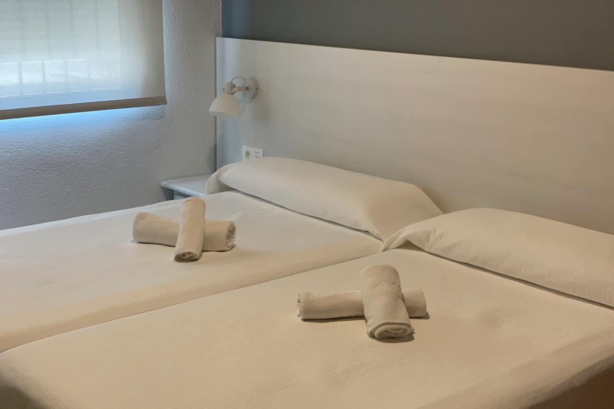 A hotel room with two beds, each with a rolled towel and a nightstand.