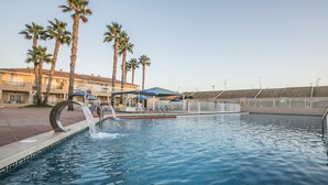 A swimming pool with palm trees, a water feature, and a building with balconies and umbrellas. - Destinos de Sol Los Alcázares (Los Alcazares)