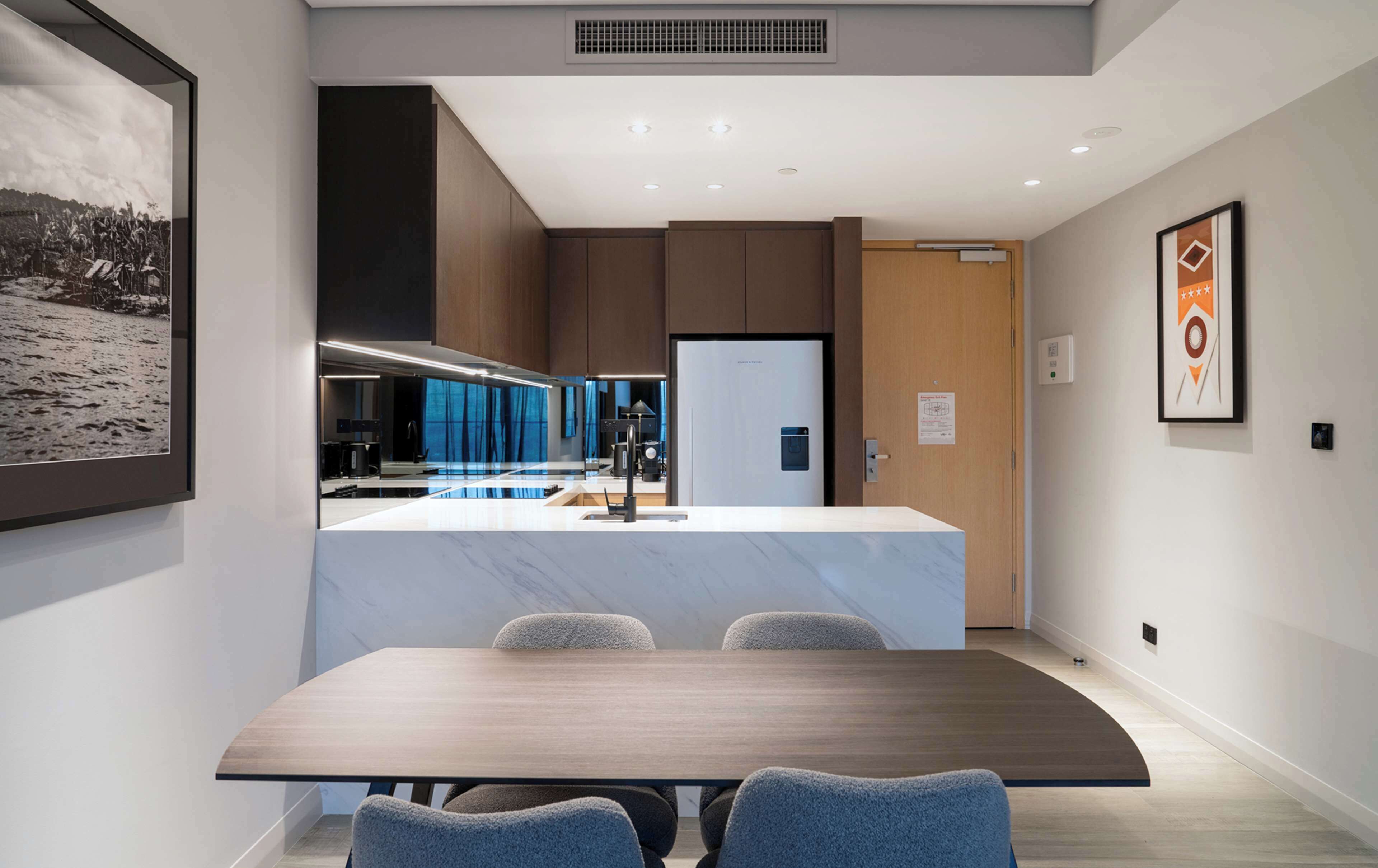 A modern kitchen with a central island, a dining area with a table and chairs, and a view of the kitchen through a glass partition.