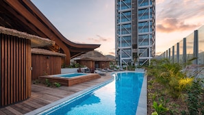 A swimming pool with wooden deck and lounge chairs, surrounded by tropical plants and a modern building with a glass facade.