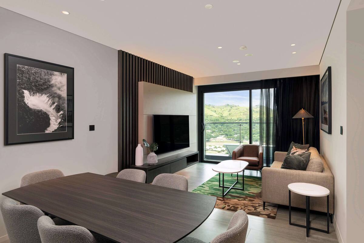 Een moderne woonkamer met een grote flatscreen televisie, een eettafel met stoelen, een bank met kussens, en een uitzicht op een groen landschap door grote ramen.