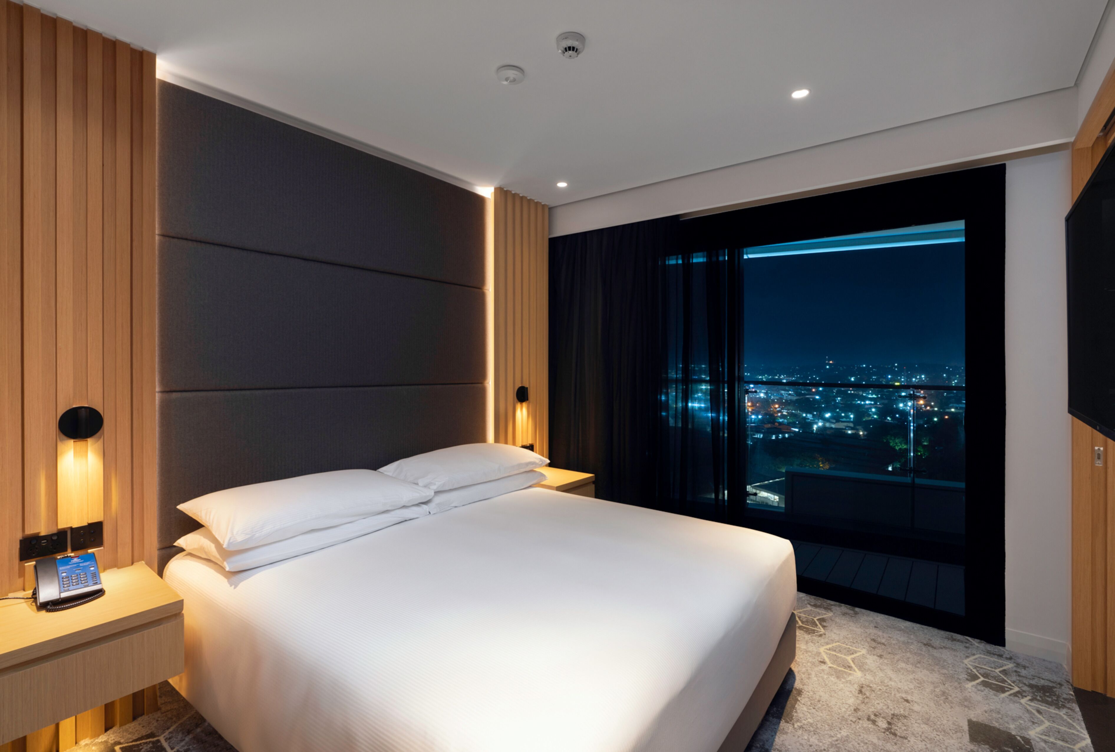 Ein modernes Hotelzimmer mit einem großen Bett, einem Nachttisch mit Telefon und Blick auf die nächtliche Stadt.