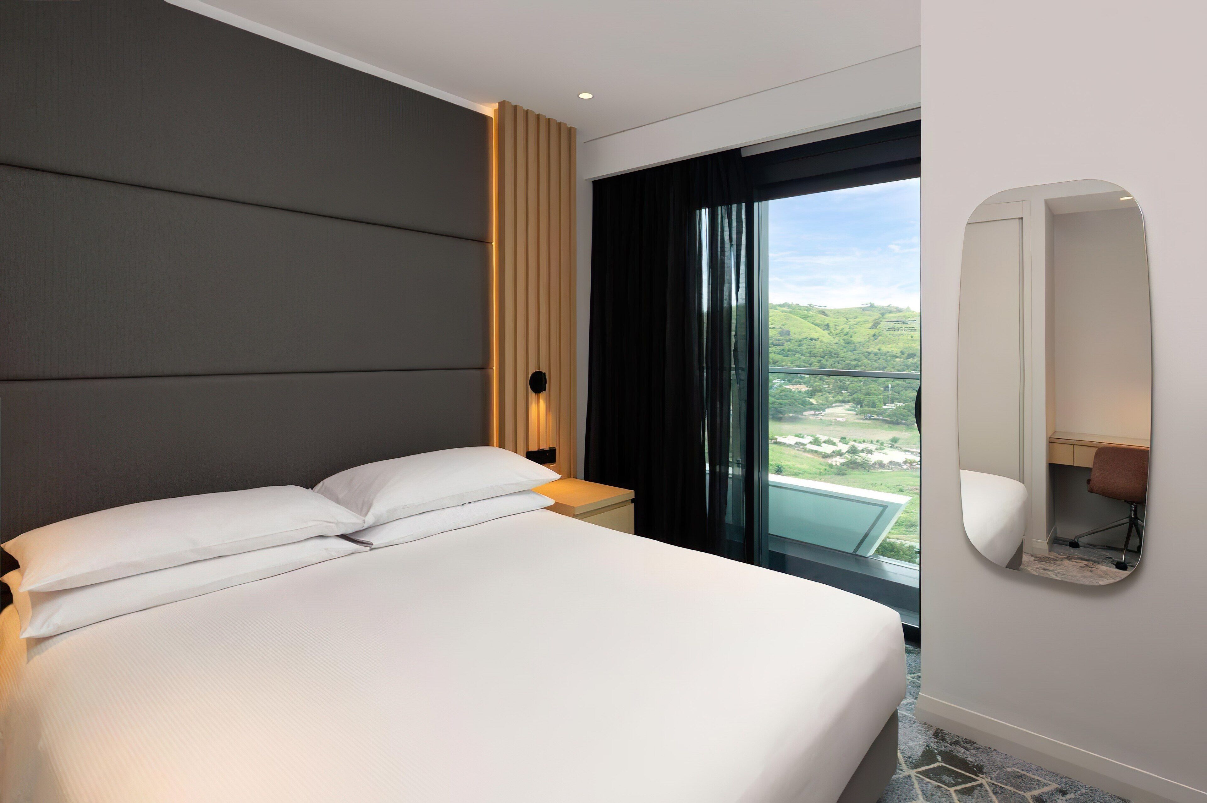 A hotel room with a large bed, a nightstand, a desk with a chair, a large window with a view, and a wall-mounted mirror.