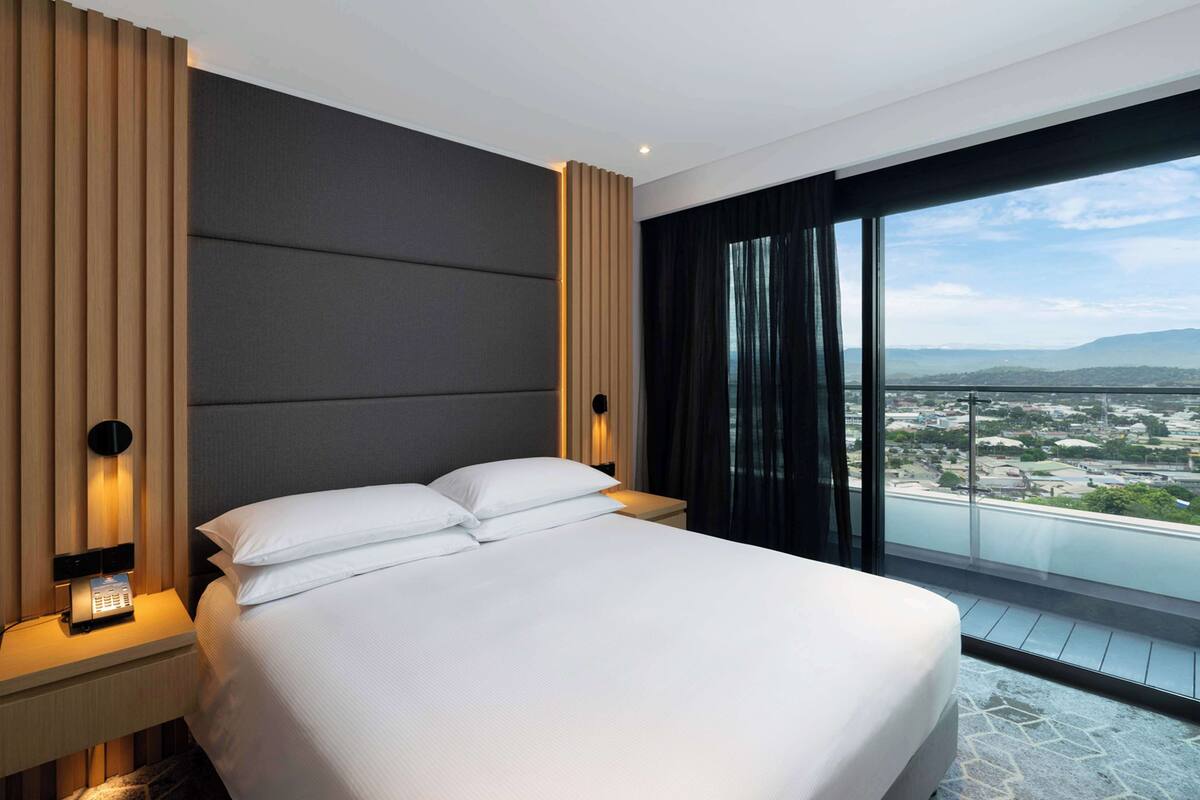 Een moderne hotelkamer met een groot bed, een nachtkastje met lamp, een bedieningspaneel aan de muur en een groot raam met uitzicht op de stad.