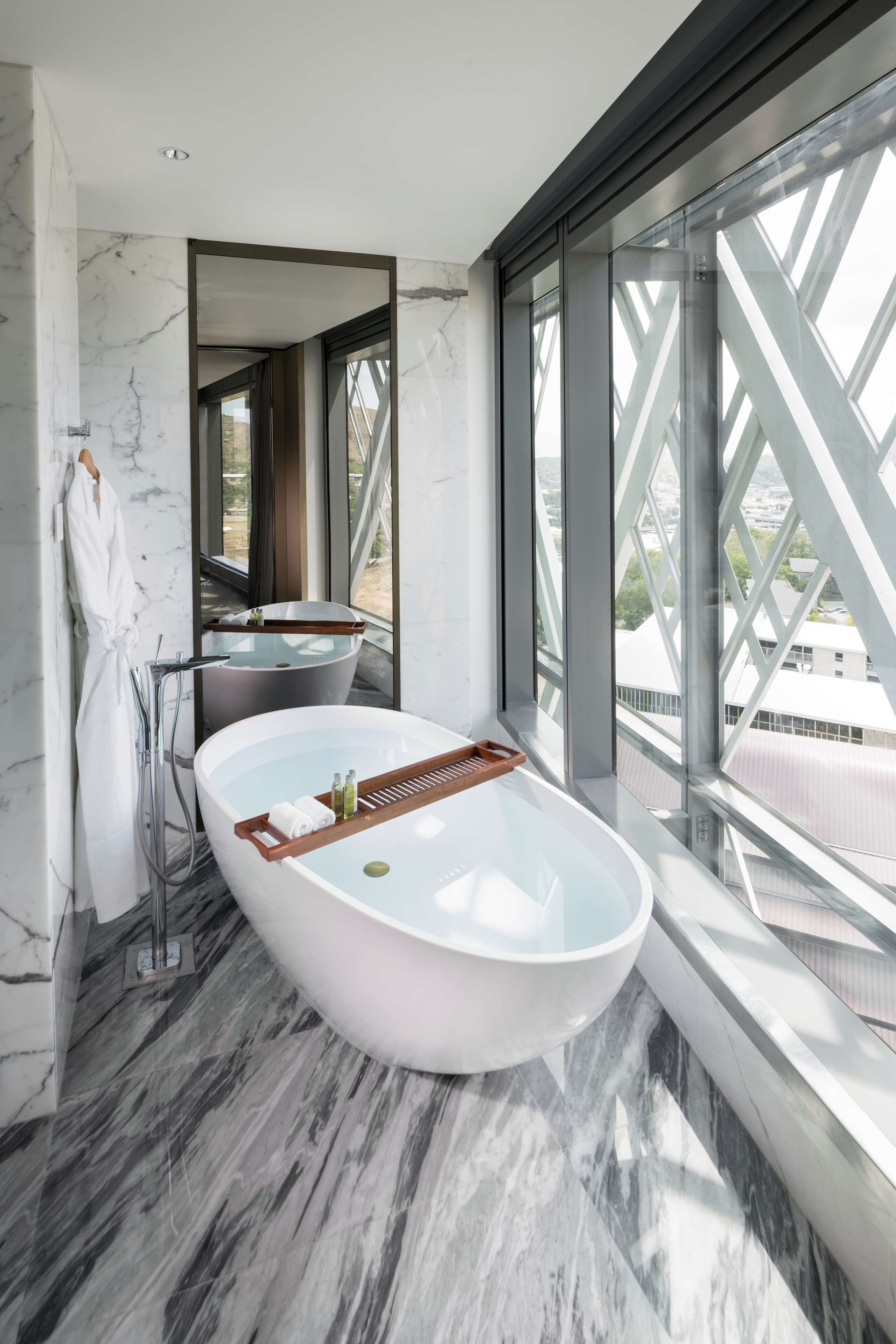 A modern bathroom with a freestanding bathtub, a glass-enclosed shower, and a large window offering a city view.