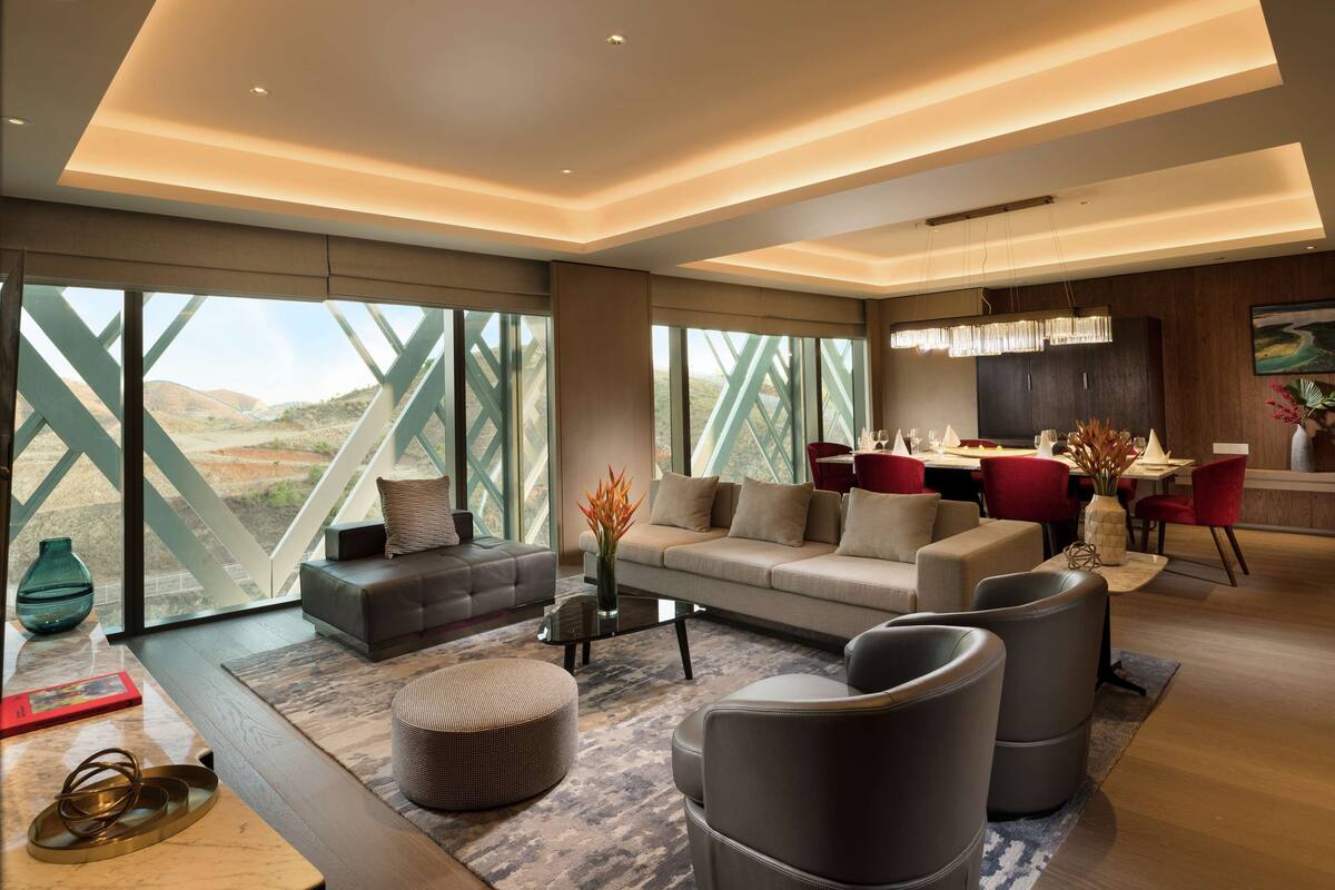Een moderne woonkamer met een bank, voetenbankje en fauteuils, een eetgedeelte met een rode tafel en stoelen, en grote ramen met uitzicht op het landschap.