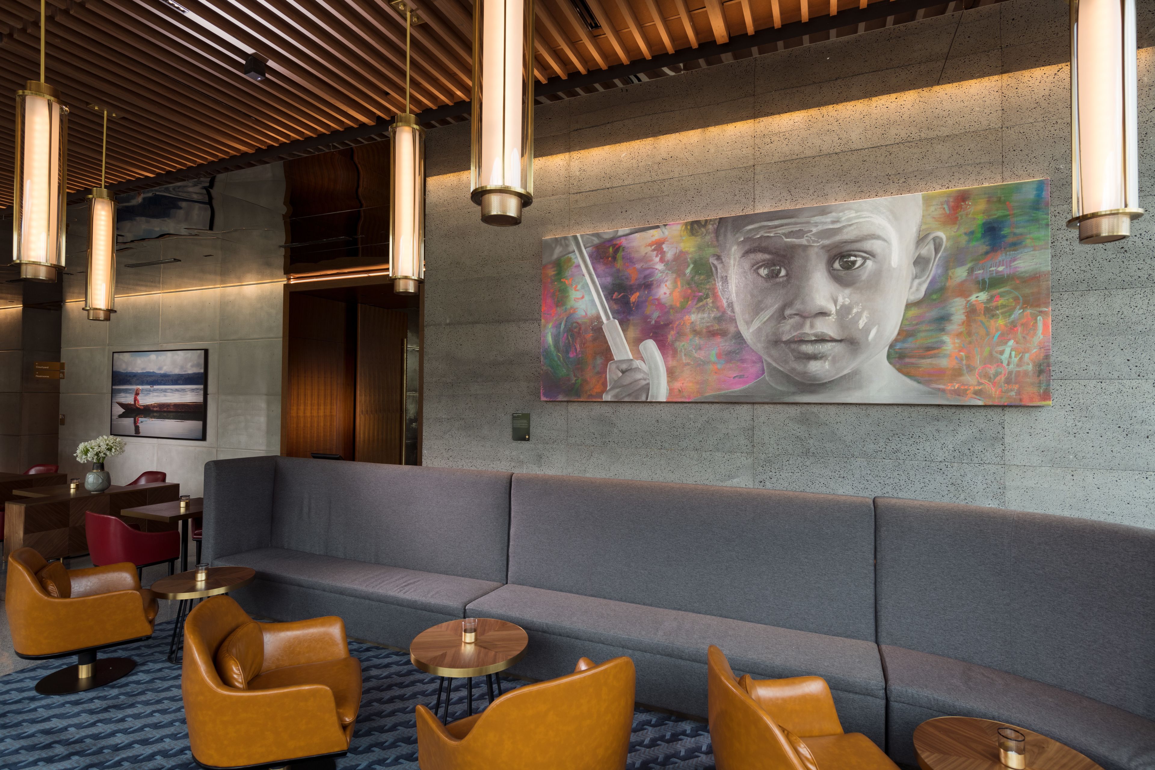 A modern lounge area with a grey sofa, leather armchairs, and round wooden tables. A large abstract painting is displayed on the wall.