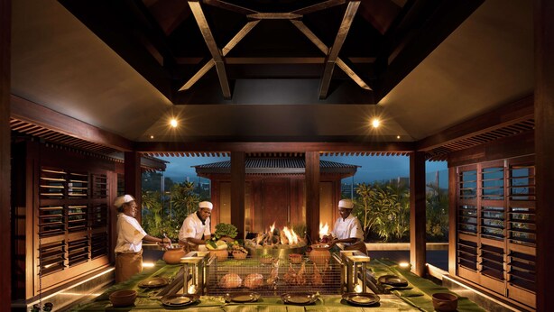 A traditional kitchen with chefs preparing food, a stone counter, and a fire pit.