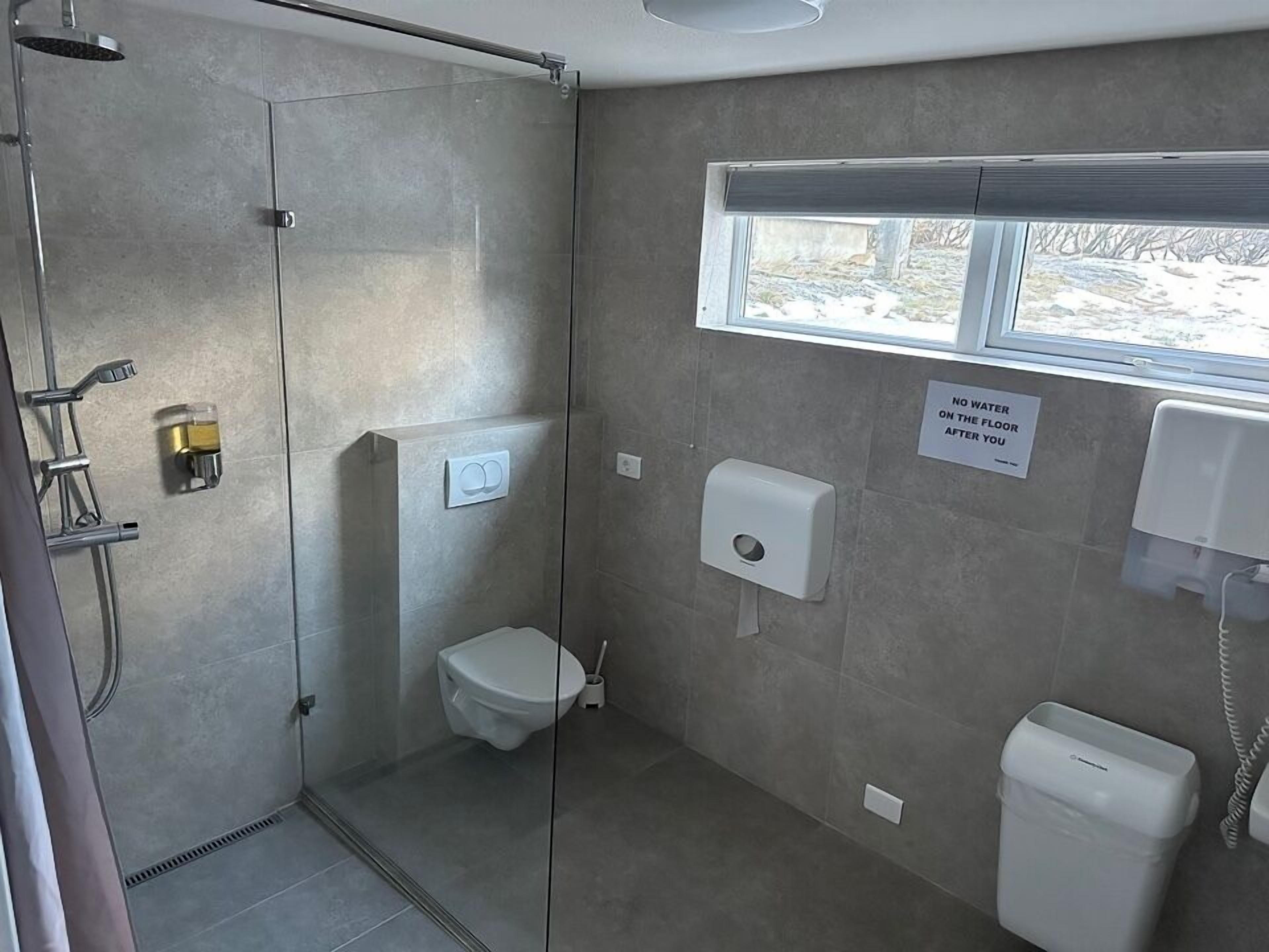 A modern bathroom with a shower, toilet, and hand dryer. A sign on the wall states "NO WATER AT THE FLOOR AFTER YOU."