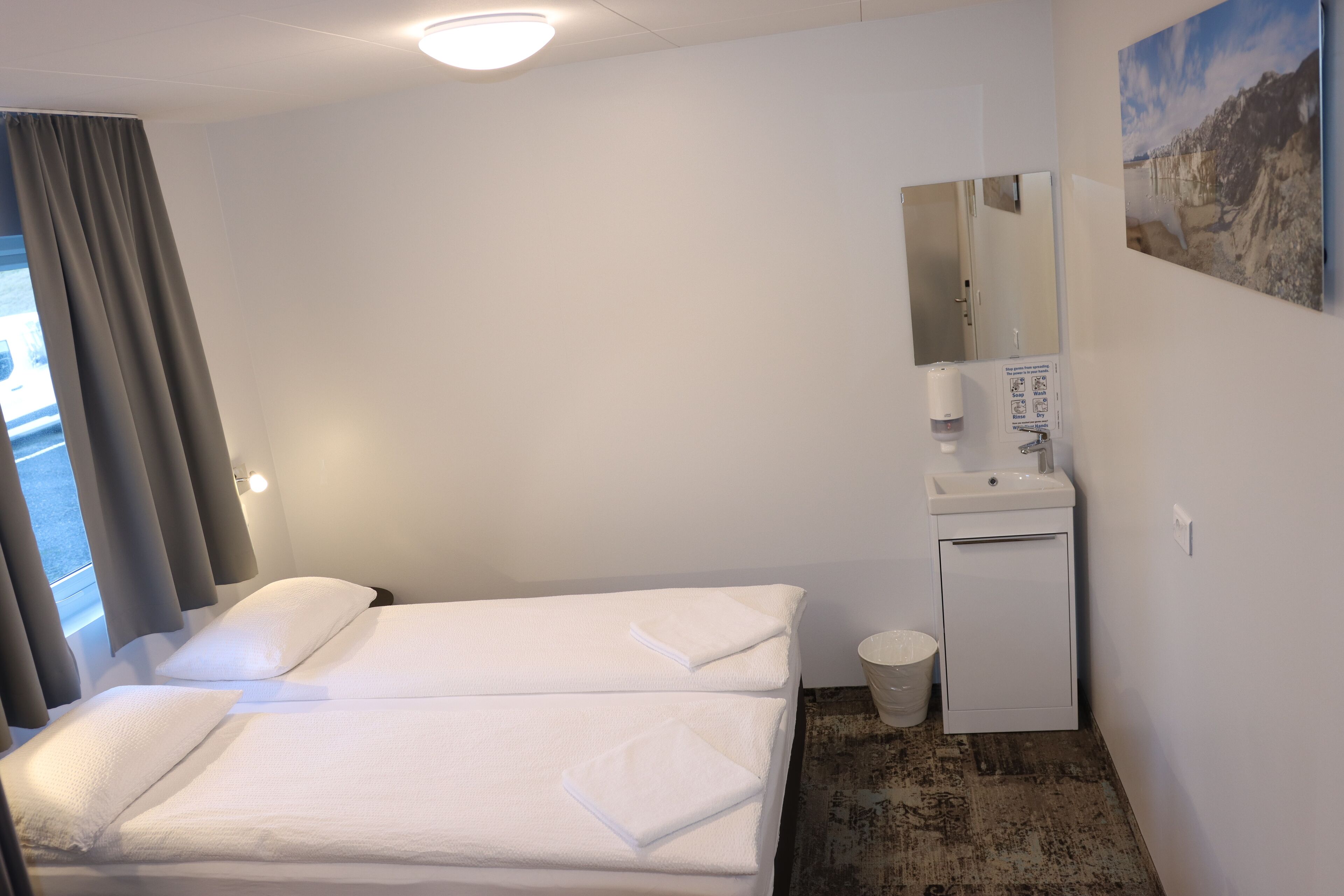 Habitación de hotel con dos camas, un lavamanos pequeño, un espejo y una ventana con cortinas.