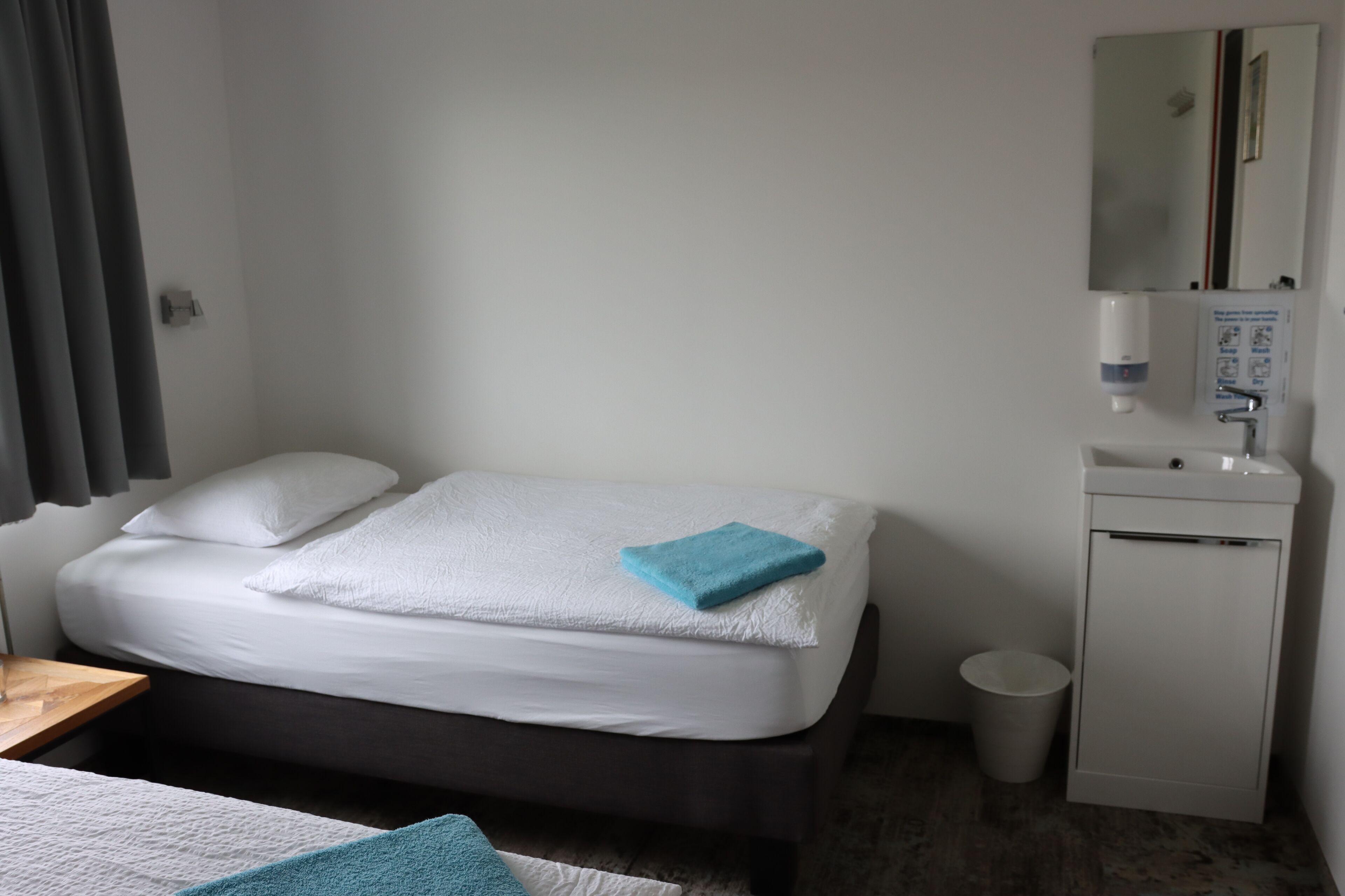 A hotel room with a bed, a small bedside table, a mirror, a sink, and a trash bin.