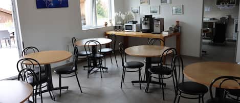 Un comedor con mesas redondas y sillas negras, una cafetera y vistas al exterior a través de una ventana.
