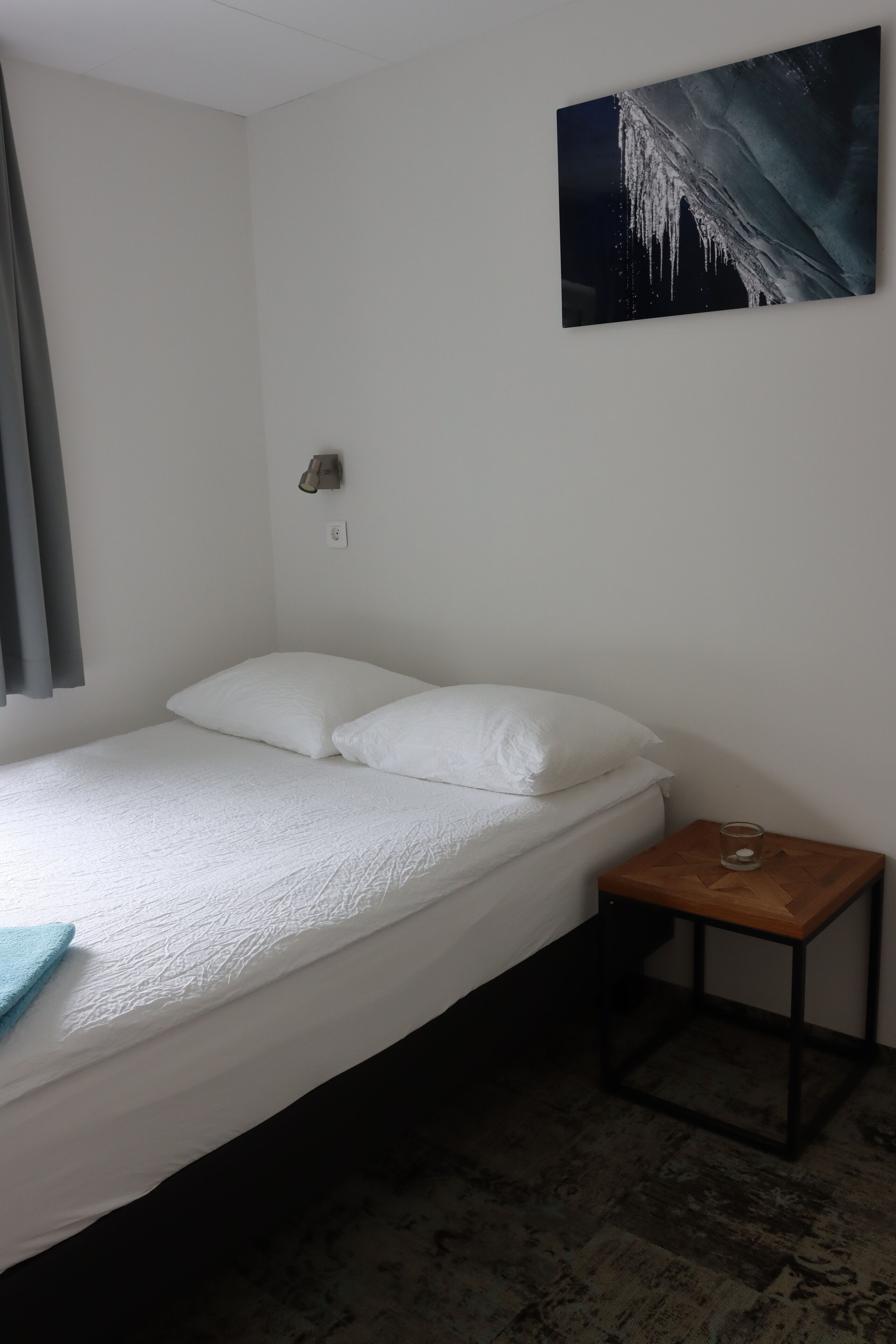 Ein Einzelbett mit weißer Bettwäsche, ein kleiner Nachttisch mit einem Glas und eine Wandleuchte.