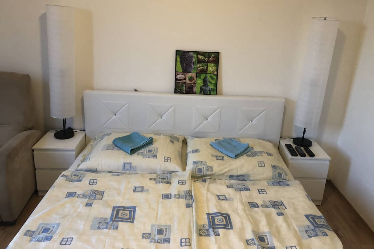 A neatly made bed with patterned bedding, two bedside lamps, a small table with a remote control, and a wall-mounted artwork.