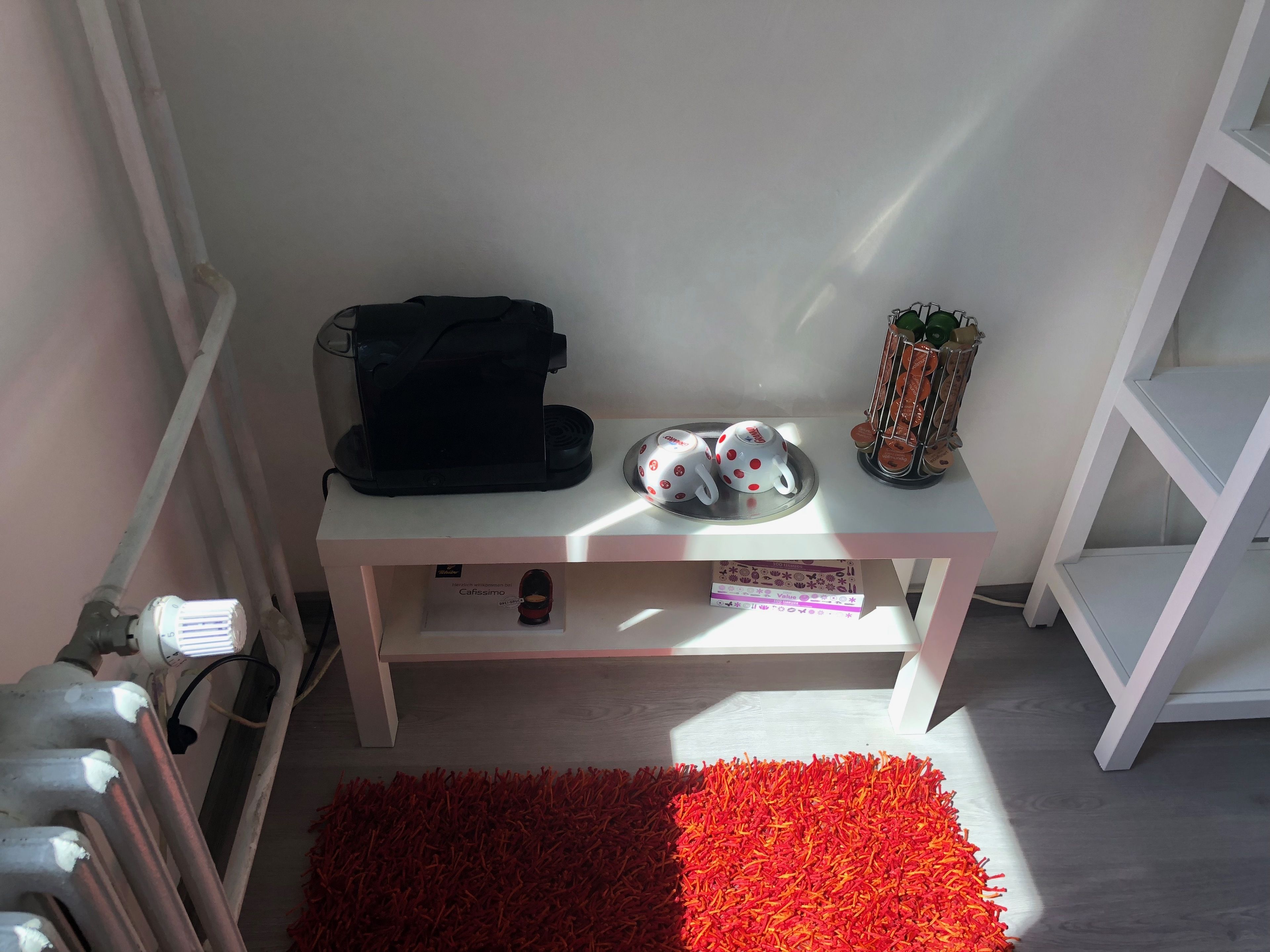 A small, neatly organized room with a white shelf holding a black bag, two white mugs with red polka dots, and a small plant. A red rug is on the floor, and a white radiator is visible on the left.