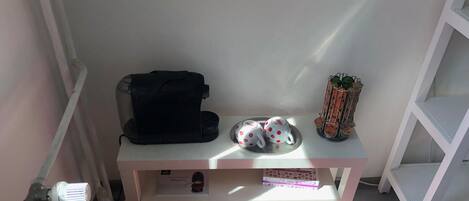A small, neatly organized room with a white shelf holding a black bag, two white mugs with red polka dots, and a small plant. A red rug is on the floor, and a white radiator is visible on the left.